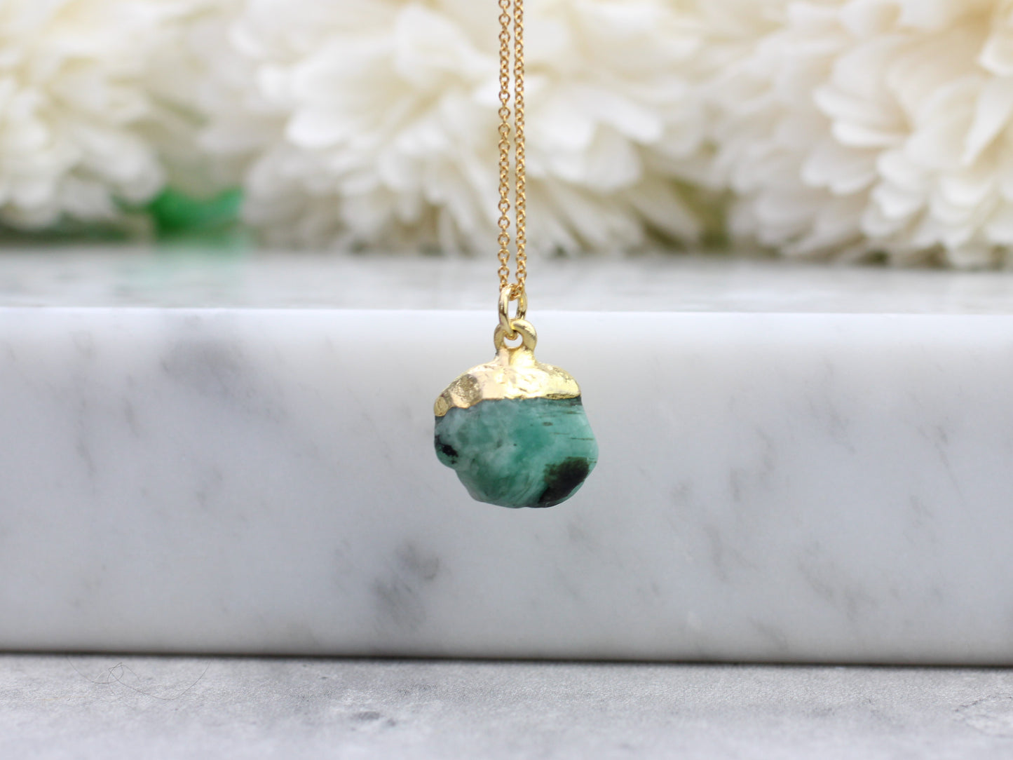 A gold-filled chain necklace with a raw emerald gemstone pendant, displayed against a floral background.