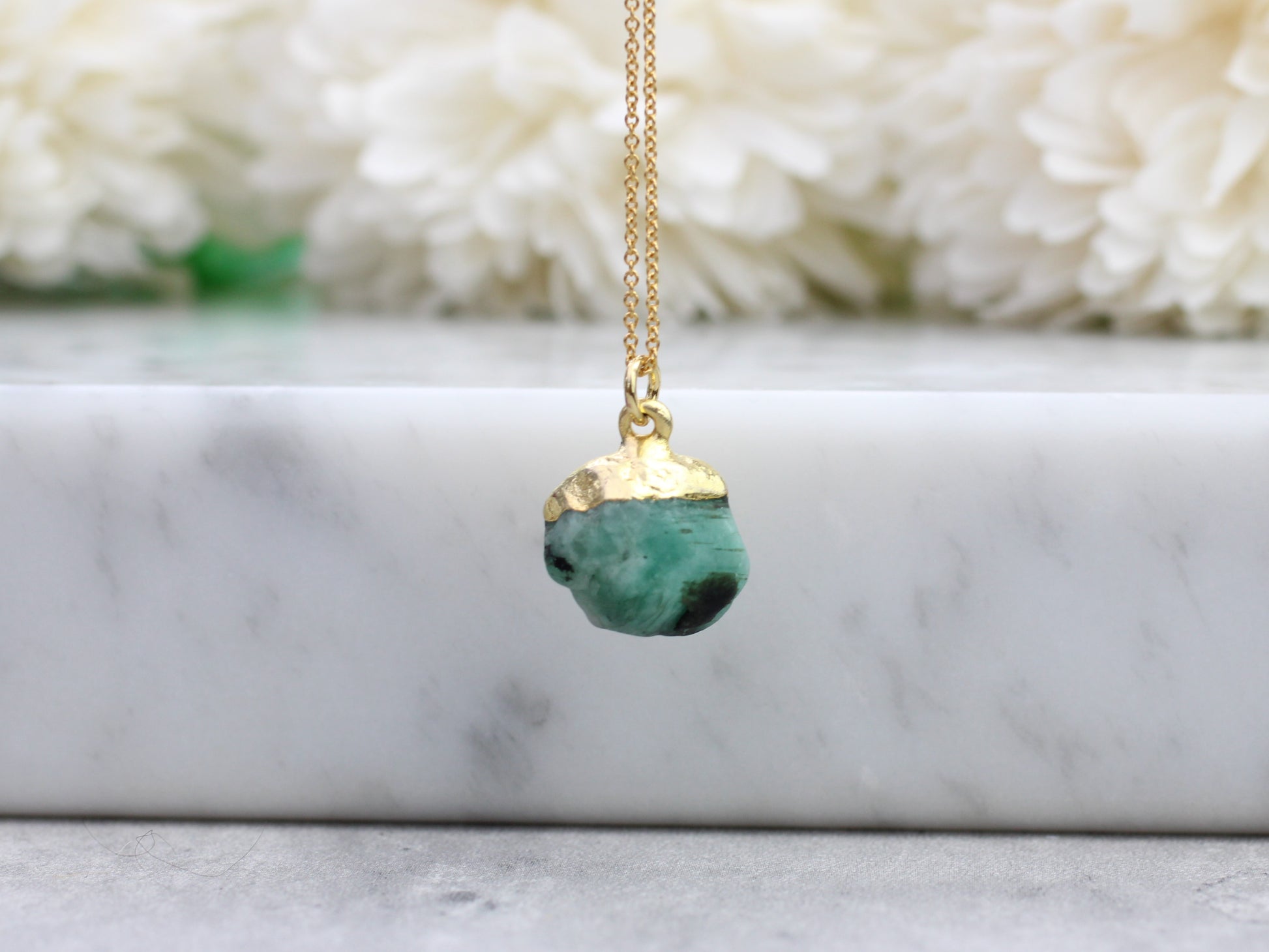 A gold-filled chain necklace with a raw emerald gemstone pendant, displayed against a floral background.