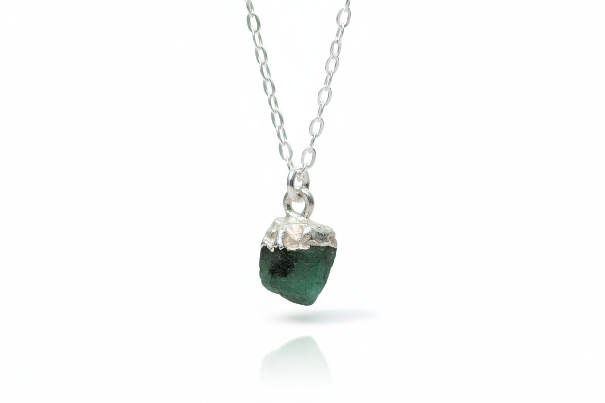 A raw emerald gemstone necklace with sterling silver electroplating, displayed against a marble background with flowers.