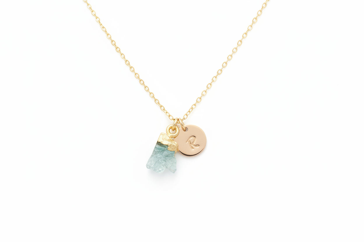 A gold necklace with a personalized initial disc and an aquamarine gemstone charm, displayed on a textured background.