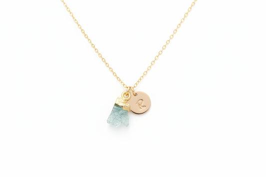 A gold necklace with a personalized initial disc and an aquamarine gemstone charm, displayed on a textured background.