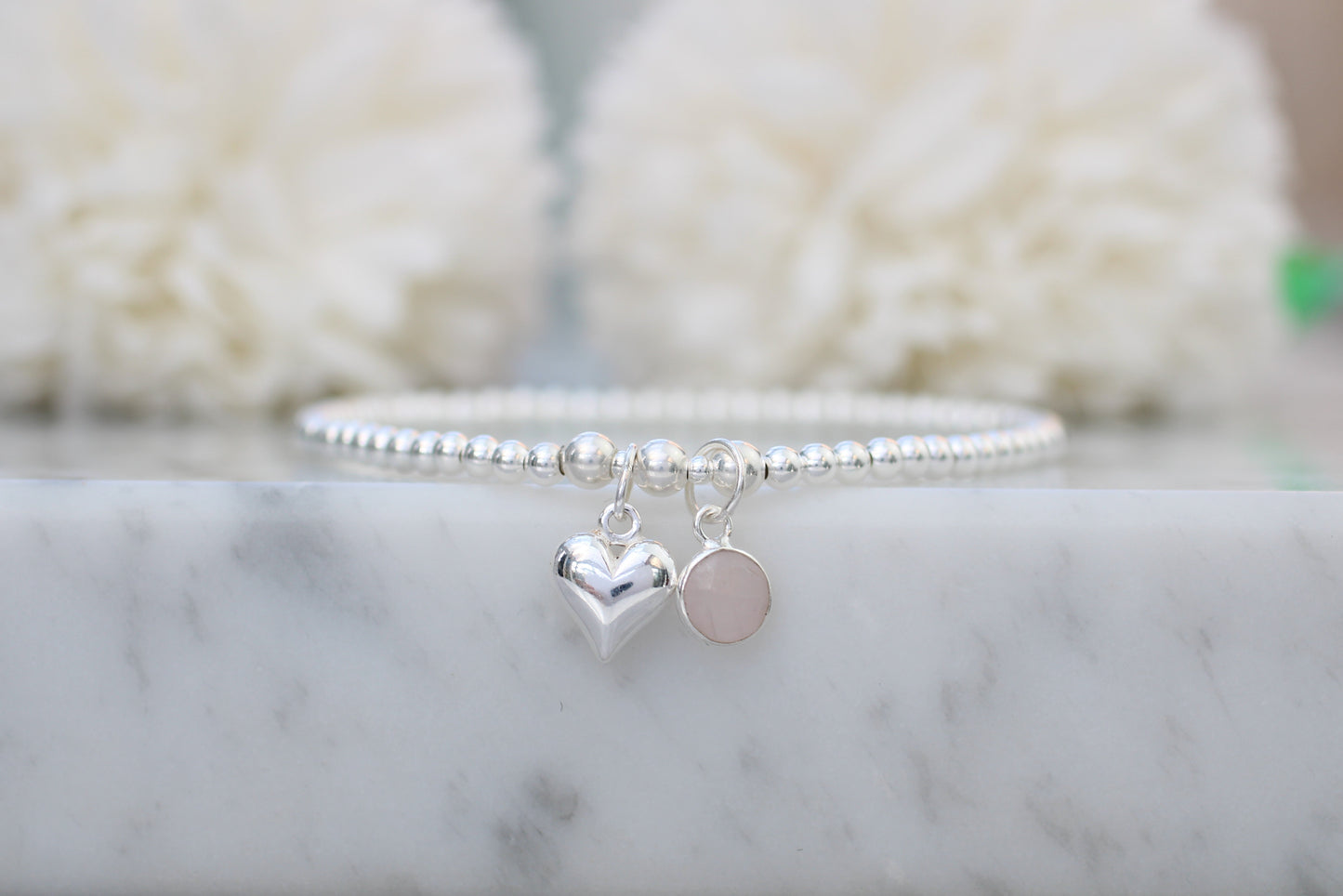 Silver bracelet with heart and round charm on a marble surface with blurred white background