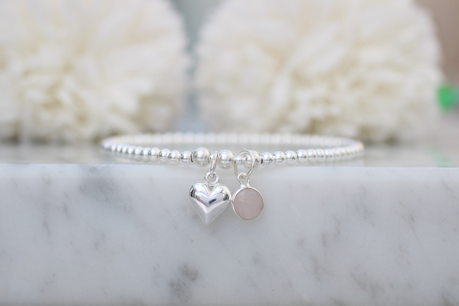 Silver bracelet with heart and round charm on a marble surface with blurred white background