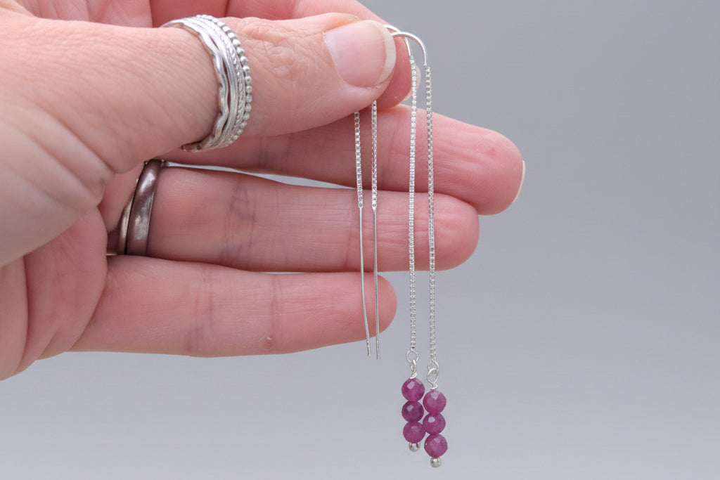 A pair of silver-colored ruby threader earrings held in a hand, with a blurred background suggesting a natural or outdoor setting.
