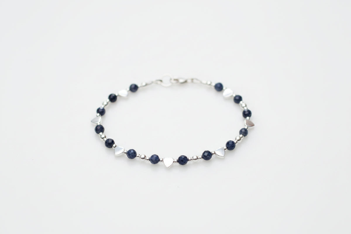 A delicate silver beaded bracelet with dark navy blue beads and small silver heart charms, displayed on a white marble surface with blurred white flowers in the background.
