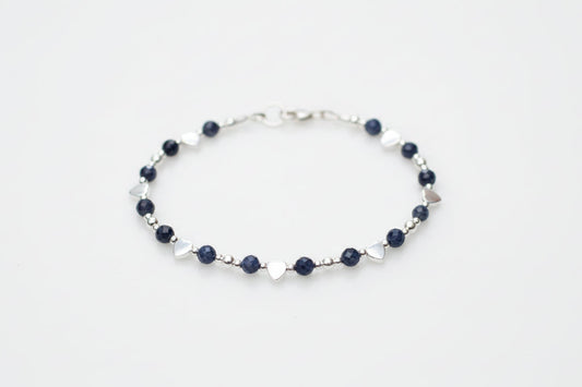 A delicate silver beaded bracelet with dark navy blue beads and small silver heart charms, displayed on a white marble surface with blurred white flowers in the background.