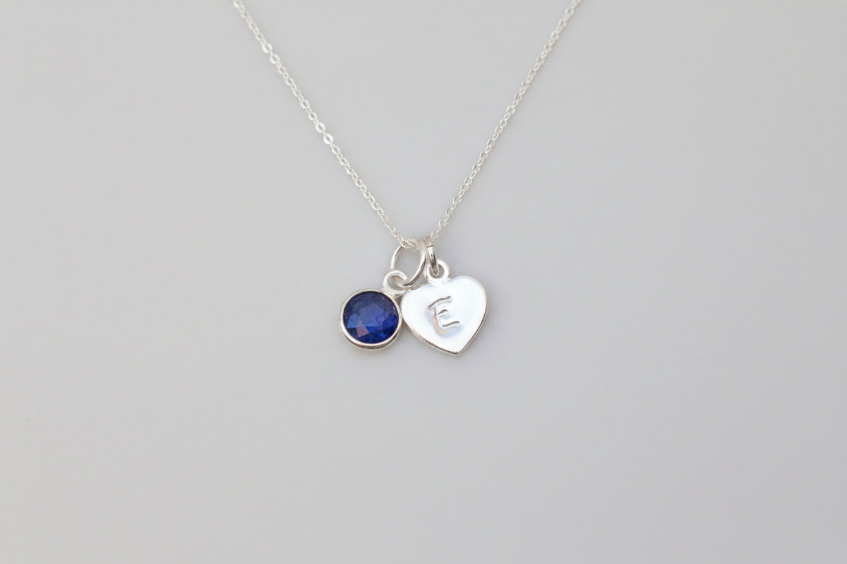 A silver necklace with two charms, one a round dark blue gemstone and the other a heart-shaped charm engraved with 'E', displayed on a white marble surface next to a white flower.