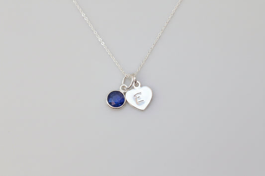 A silver necklace with two charms, one a round dark blue gemstone and the other a heart-shaped charm engraved with 'E', displayed on a white marble surface next to a white flower.