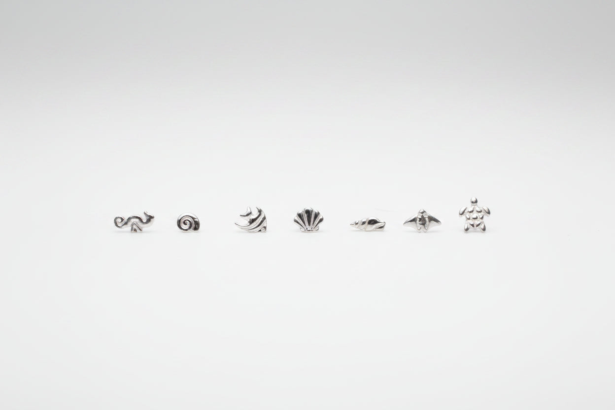 seven small silver sea-themed stud earrings arranged in a row on a white marble surface, with two large white decorative flowers blurred in the background.
