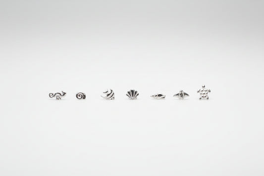 seven small silver sea-themed stud earrings arranged in a row on a white marble surface, with two large white decorative flowers blurred in the background.