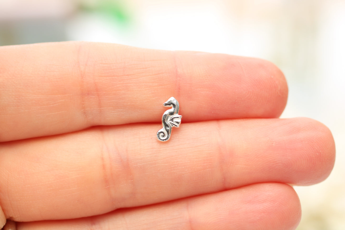 A small silver seahorse charm resting on the fingers of a hand, shown against a soft, blurred light background.