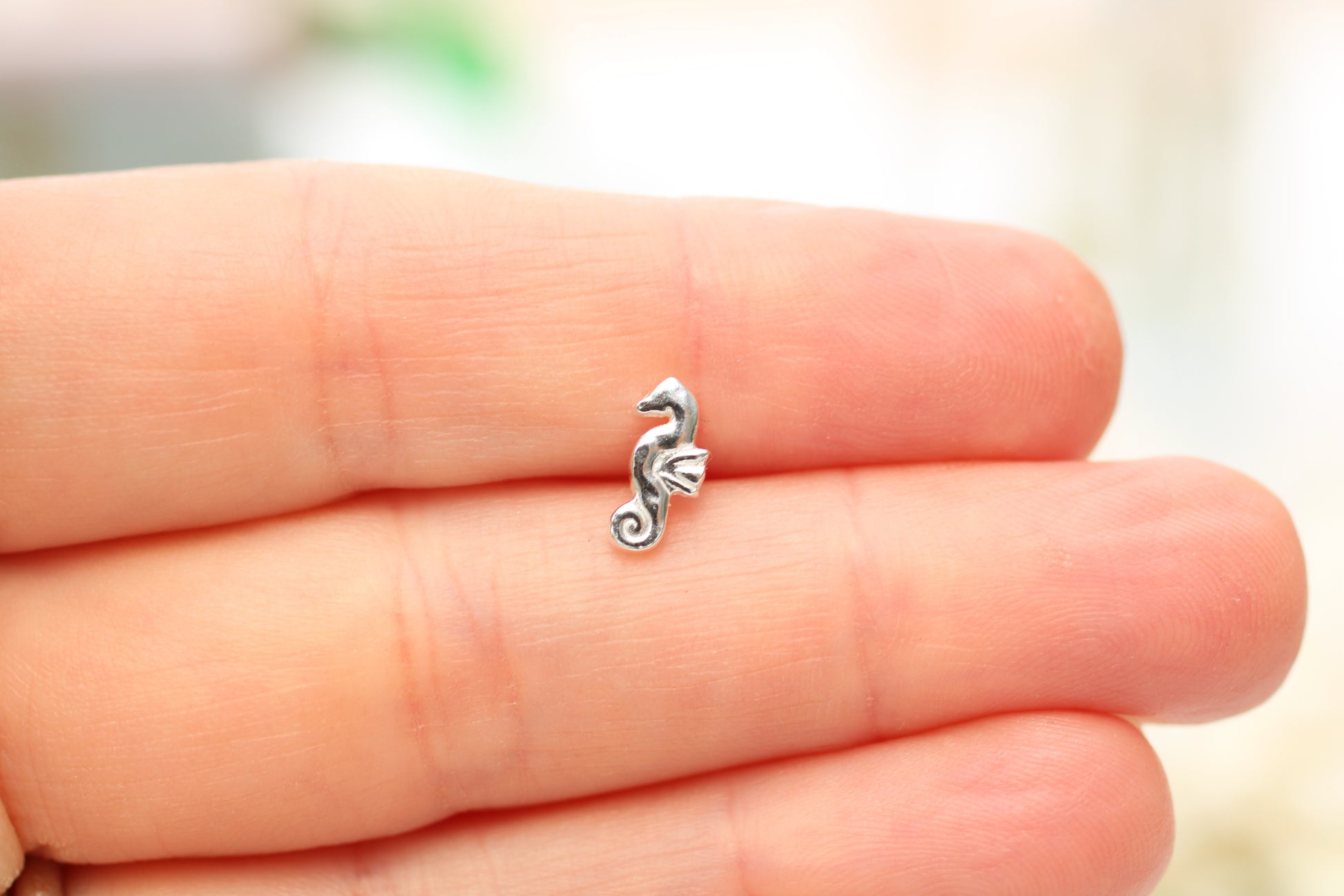 A small silver seahorse charm resting on the fingers of a hand, shown against a soft, blurred light background.