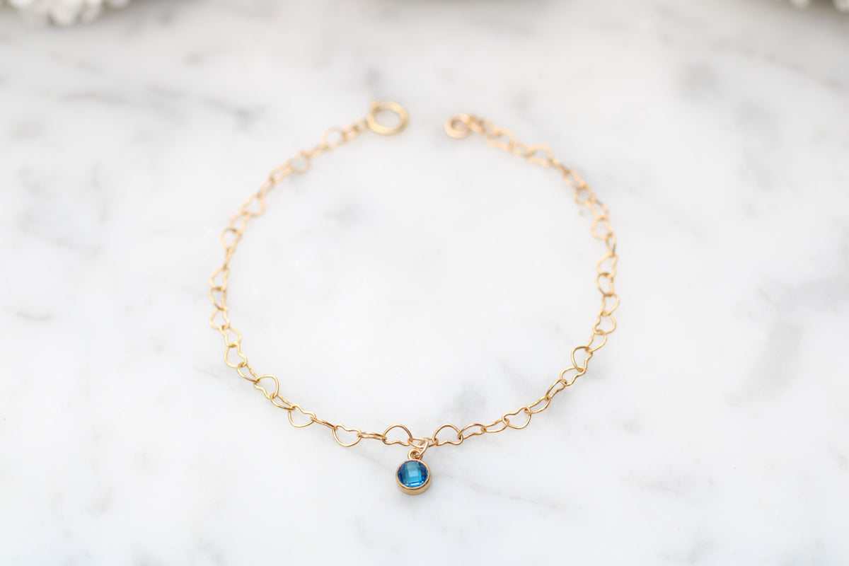 A delicate gold chain bracelet with a small oval blue gemstone pendant, displayed on a white marble surface.