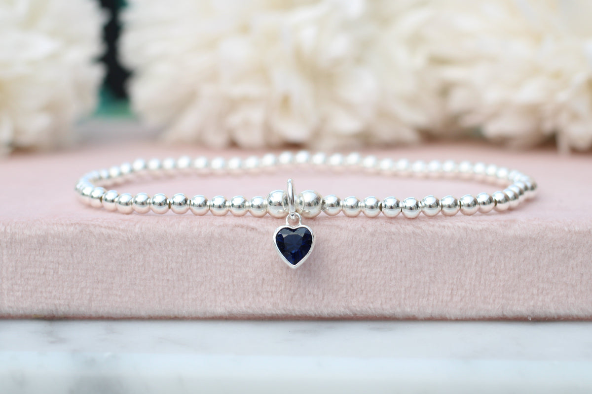 A delicate silver beaded bracelet with a small heart-shaped dark blue gemstone charm, resting on a pink textured box with soft white flowers in the background.
