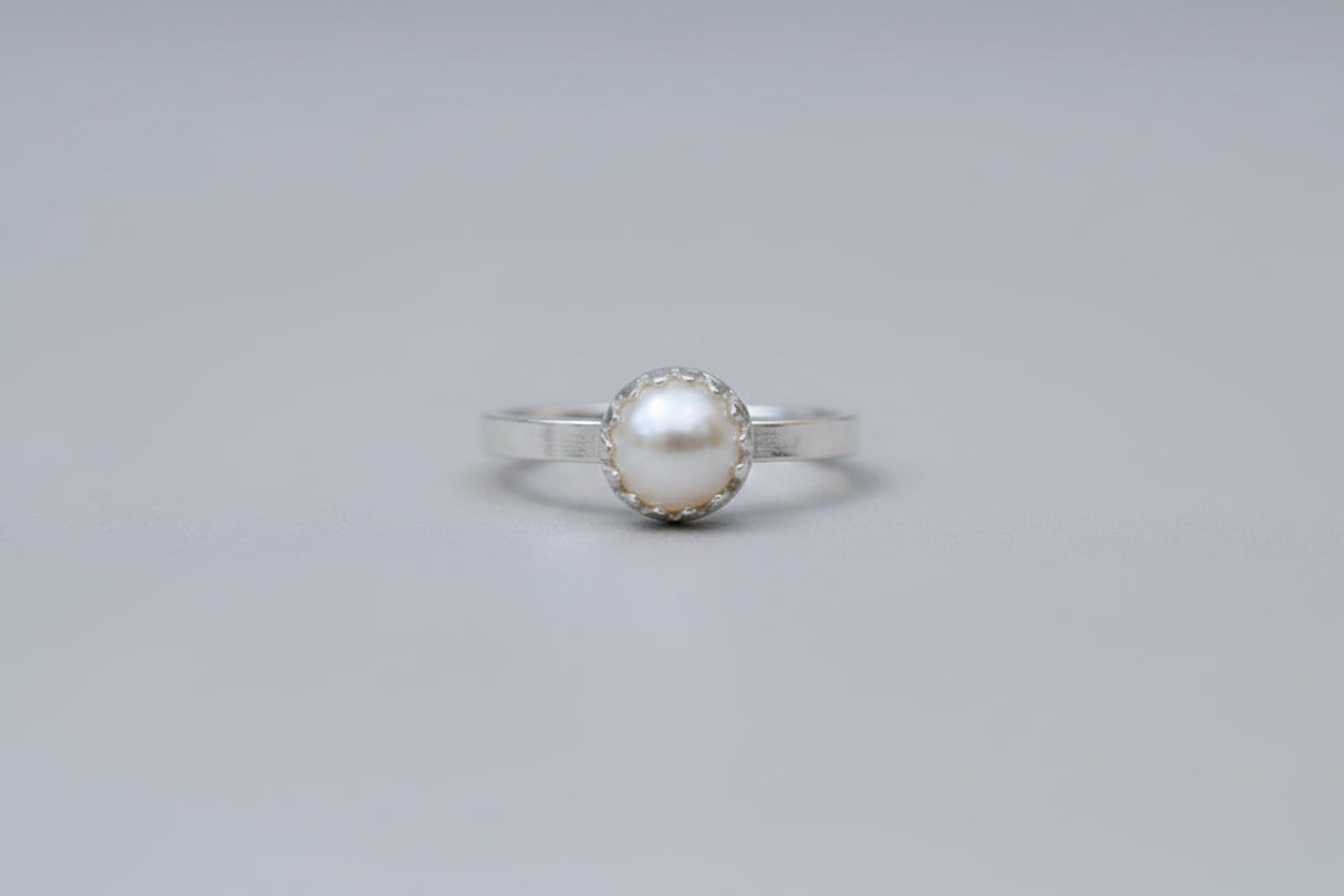 A silver ring with a round white pearl in a bezel setting, resting on a wooden surface with a blurred white flower in the background.
