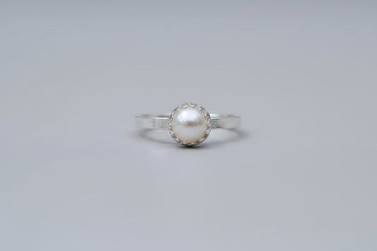 A silver ring with a round white pearl in a bezel setting, resting on a wooden surface with a blurred white flower in the background.