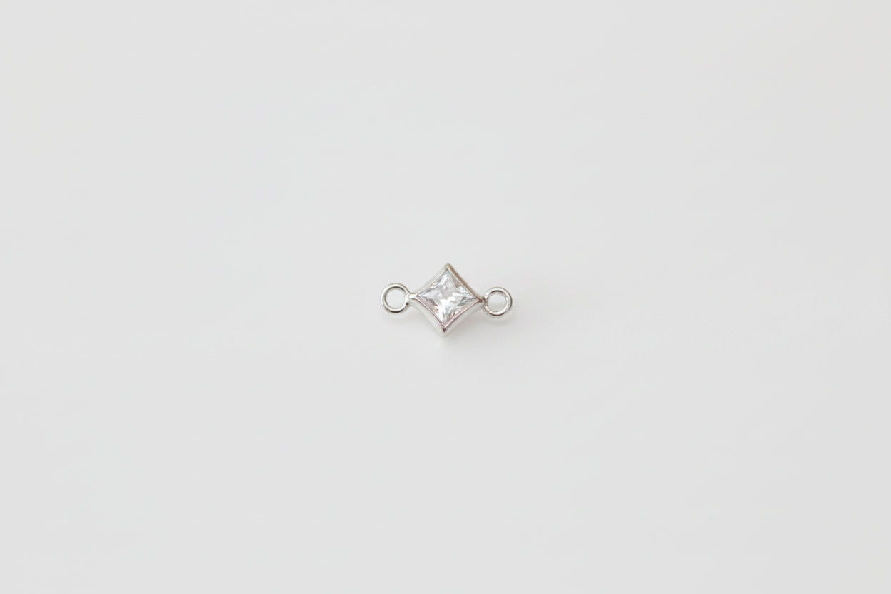 A small silver-toned jewelry charm with a clear, diamond-shaped gemstone in a bezel setting, flanked by two small loops, displayed on a light gray marble surface.