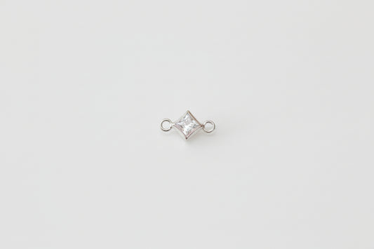 A small silver-toned jewelry charm with a clear, diamond-shaped gemstone in a bezel setting, flanked by two small loops, displayed on a light gray marble surface.