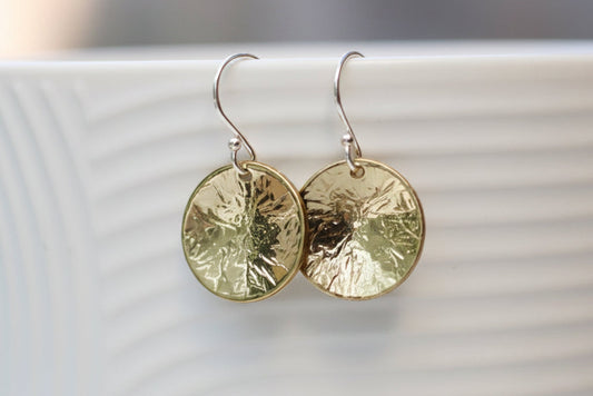 A pair of textured gold and silver dangle earrings hanging from a white ribbed ceramic mug in a softly blurred setting.