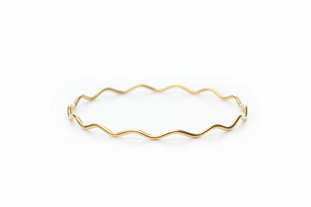 A delicate gold wavy wire bangle bracelet resting on a white marble surface with a soft-focus background of white pom-pom flowers.
