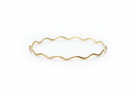A delicate gold wavy wire bangle bracelet resting on a white marble surface with a soft-focus background of white pom-pom flowers.