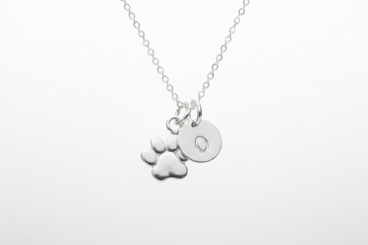 A silver necklace with a delicate chain and two charms: a paw print and a round disc engraved with the letter 'O', shown on a clean white background.