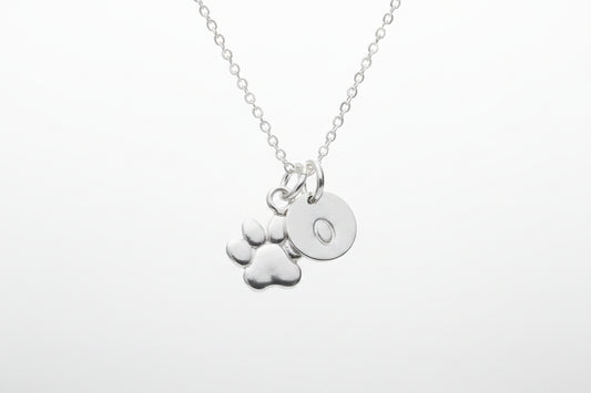A silver necklace with a delicate chain and two charms: a paw print and a round disc engraved with the letter 'O', shown on a clean white background.